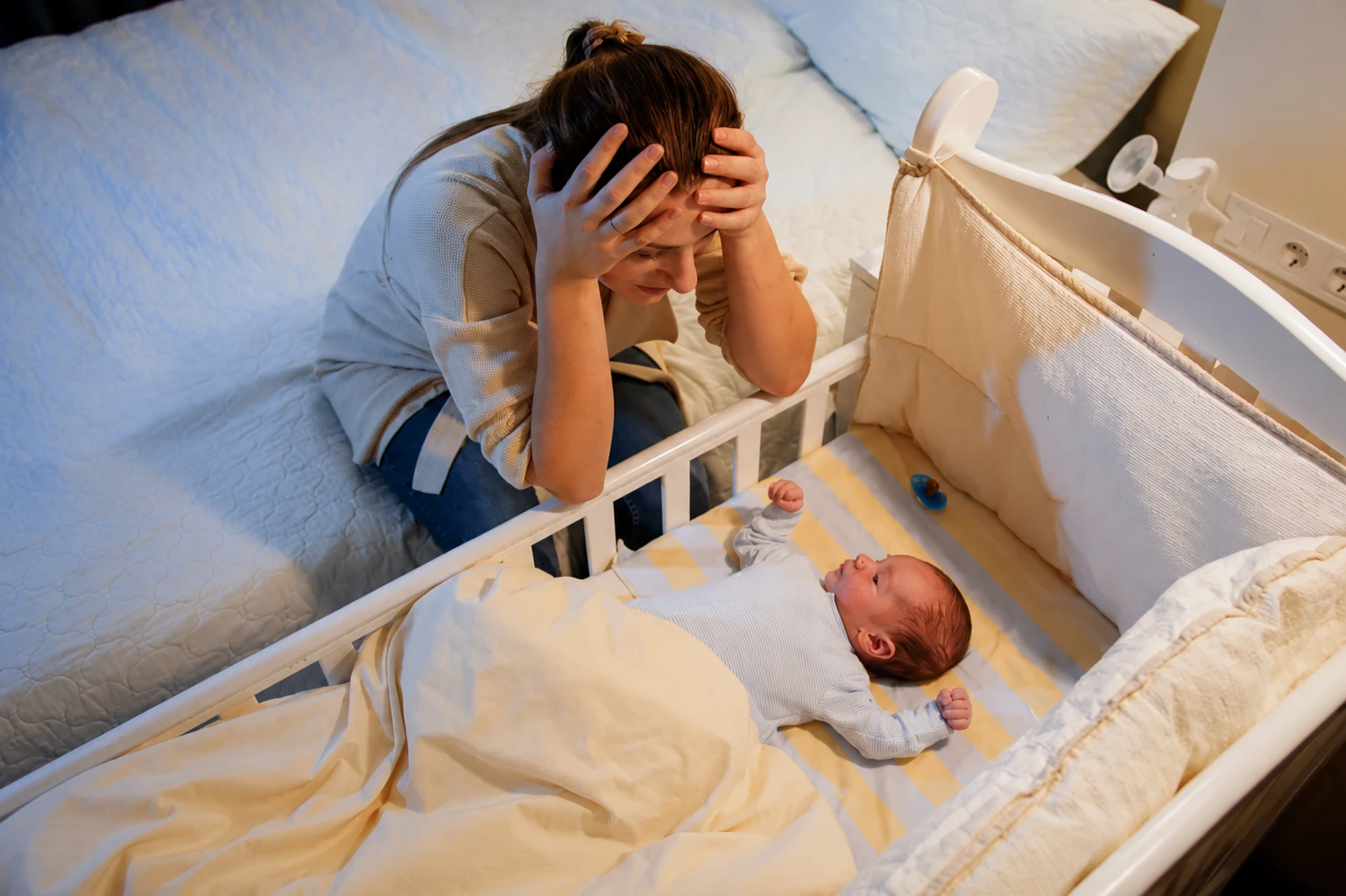 Madre agotada con las manos en la cabeza junto a la cuna con el bebé despierto de madrugada